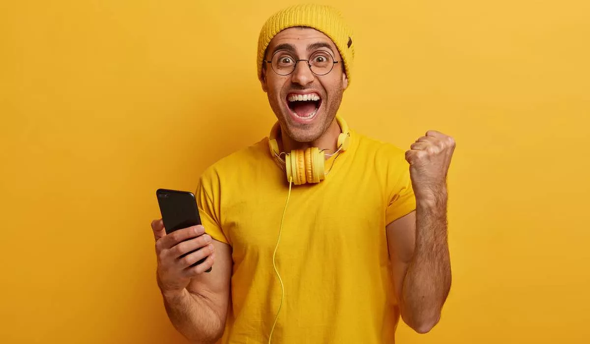 el hombre alegre aplaude y celebra la victoria del juego del telefono inteligente levanta el puno se regocija al terminar el nivel dificil usa ropa elegante y vivida